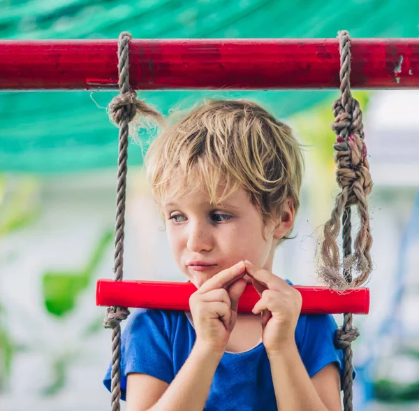 Close portrait Sad lonely freckled blue eyed cute blond little boy in t-shirt sitting on a rope ladder outside on playground in kindergarten. Daycare, activity, childhood, friendship, relationship — Stock Photo, Image