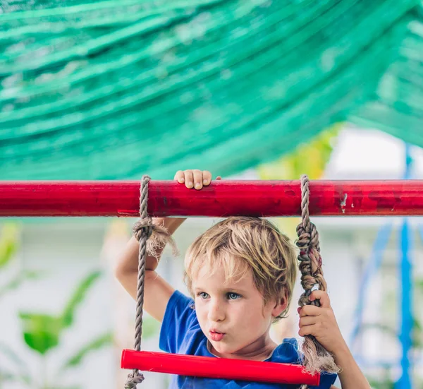 Close portrait Happy smile freckle blue eyed cute blond little boy sitting on a rope ladder, playing outside on playground in kindergarten. Daycare, activity, simple joys of childhood, summer — Stock Photo, Image