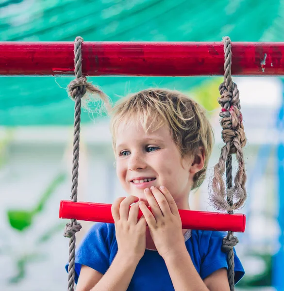 Potret dekat Senyum gembira mata biru bermata biru anak kecil pirang lucu duduk di tangga tali, bermain di luar di taman bermain di TK. penitipan anak, aktivitas, sukacita sederhana masa kecil, musim panas — Stok Foto