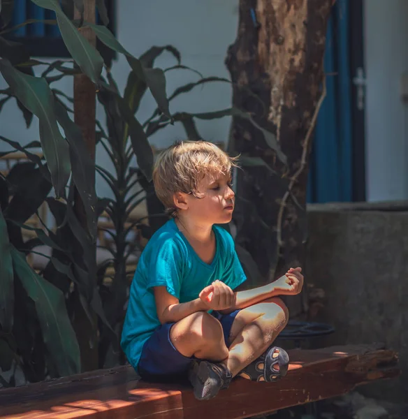 Calm little male boy sit on couch outside garden practice yoga. Young generation modern child rest relieve negative emotions with closed eyes in lotus pose at sunrise. Mindfulness meditation concept