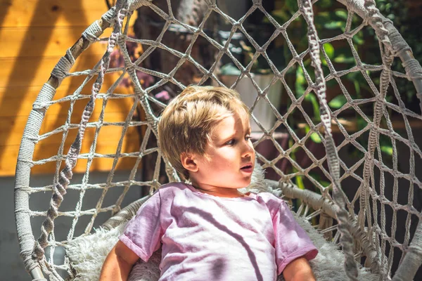 Cute smiling freckled blond boy emotion facial expressions gestures, intriguing funny mischievous in pink, look sun beams sit Hammock Chair Hanging. Happy childhood mood, children behaviour concept — Stock Photo, Image