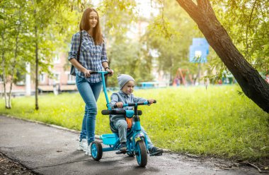 Genç anne, yürümeye yeni başlamış bir çocukla bir çocuğun bisikletini itiyor. Bisiklet sürmeyi öğrenme ve ailenle eğlenme kavramı..