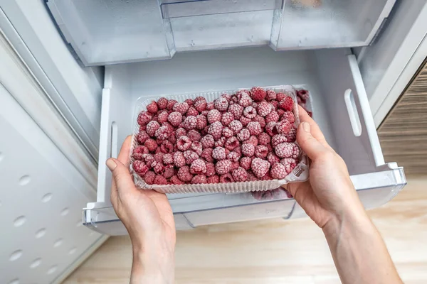 A person is taking a container of frozen raspberries out of the freezer of the fridge. Concept of frozen food, long term storage products.