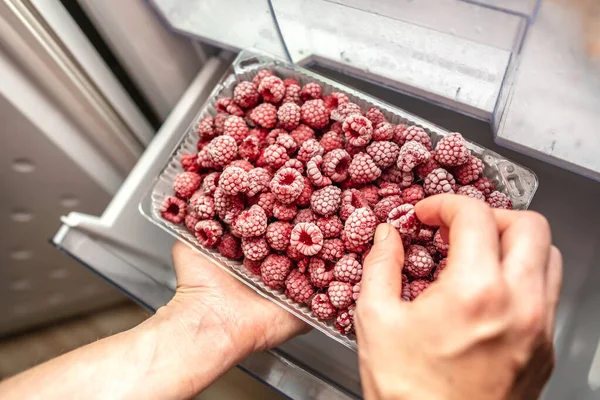 A person is taking a container of frozen raspberries out of the freezer of the fridge. Concept of frozen food, long term storage products.
