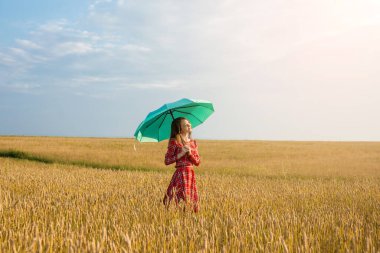 Kırmızı elbiseli genç bir kadın ve elinde buğday tarlasında altın dikenlerin arasında yeşil bir şemsiye tutuyor. Koruma, sakinlik ve özgürlük kavramı.
