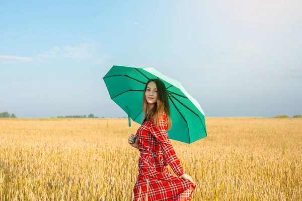 身穿红色长裙 头戴绿色雨伞的年轻女子正在麦田里散步 享受着温暖的秋日 清新的空气和自由 — 图库照片