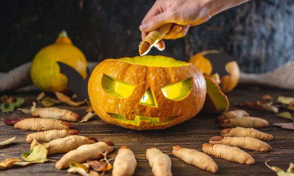 Hand is putting the cookie in the form of a finger inside the pumpkin with the face cut out. Concept of celebrating Halloween and the sinister atmosphere.