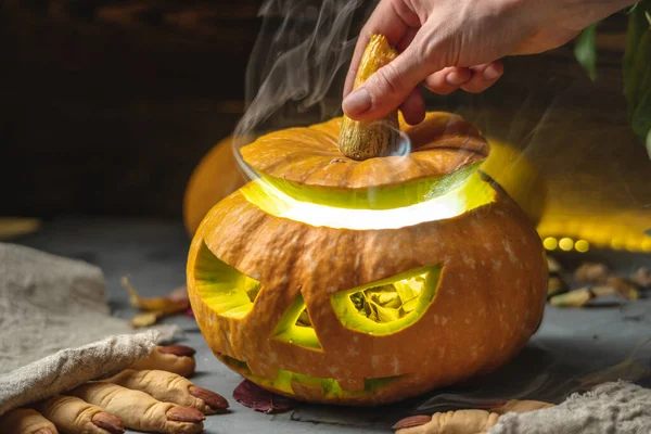 Hand is opening the pumpkin with the face cut out and smoke comes out of it. Concept of celebrating Halloween and a magical mysterious atmosphere.