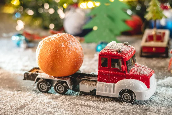 Red and white toy truck is going along a snow covered road against a background of festive lights and pines and carrying a tangerine. Concept of holiday mood and preparation for the celebration.