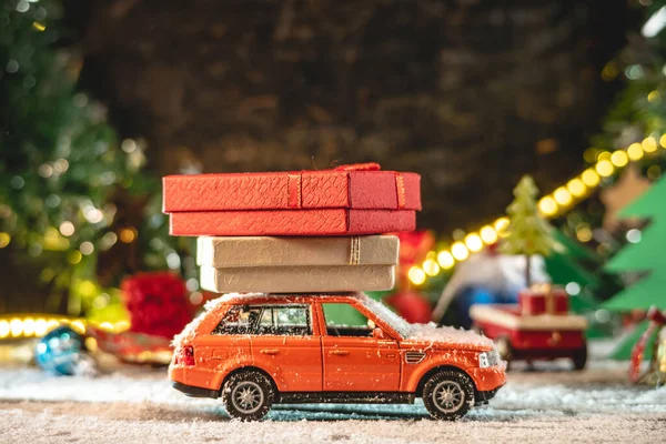 An orange toy car is going along a snow covered road against the background of festive lights and pines and carrying boxes with gifts. Concept of Christmas mood and preparation for the celebration.