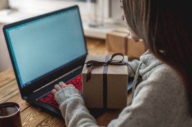 Genç bir kadın dizüstü bilgisayarın önünde oturuyor, onu kullanıyor ve bir hediye kutusu tutuyor. Çevrimiçi hediye alma ve seçme kavramı.