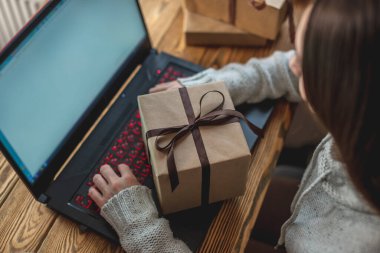 Genç bir kadın dizüstü bilgisayarın önünde oturuyor, onu kullanıyor ve bir hediye kutusu tutuyor. Çevrimiçi hediye alma ve seçme kavramı.