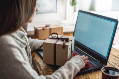 Genç bir kadın dizüstü bilgisayarın önünde oturuyor, onu kullanıyor ve bir hediye kutusu tutuyor. Çevrimiçi hediye alma ve seçme kavramı.