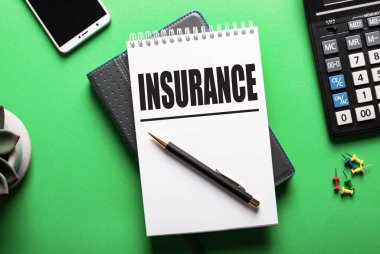 INSURANCE kelimesi, hesap makinesi ve telefonun yanındaki yeşil bir masanın üzerine beyaz bir deftere yazılmıştır. Sigorta konsepti