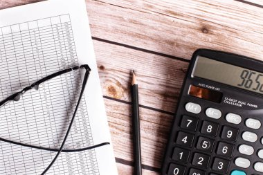 Karanlık bir ahşap masada bir hesap makinesi, gözlük, bir kalem ve bir belge yatıyor. Üst görünüm kapalı. İş konsepti