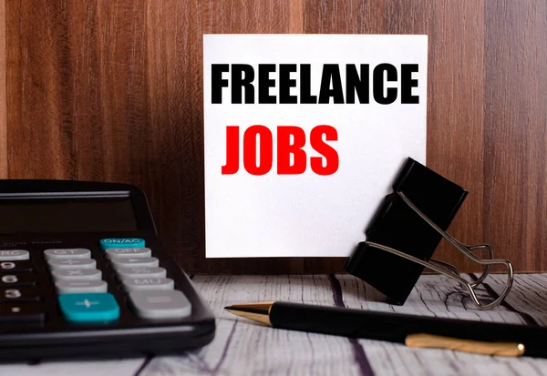 FREELANCE JOBS written on a white card next to a calculator and a pen on a wooden background. Business concept