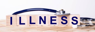 Üzerinde ILLNESS yazan tahta küpler ve tahta bir arka planda steteskop var. Tıbbi konsept
