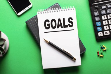 Yeşil bir arkaplanda bir telefon, bir hesap makinesi ve içinde GOALS sözcüğünün yazılı olduğu bir günlük bulunur.