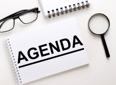 AGENDA kelimesi defter, siyah çerçeveli gözlük ve büyüteç gibi arka planda beyaz bir deftere yazılmıştır..