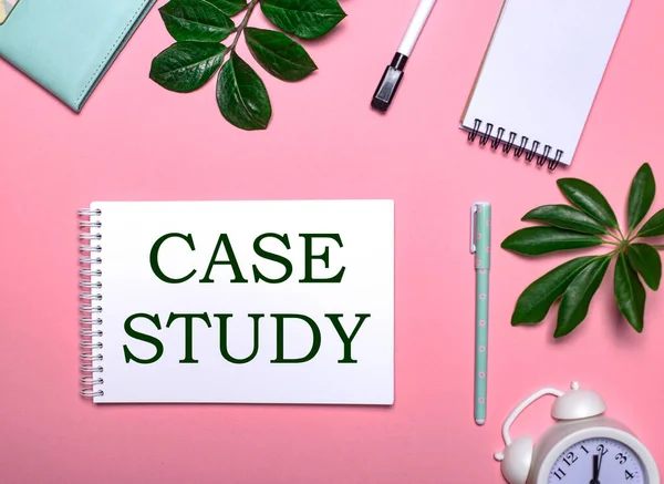 CASE STUDY is written in green on a white notepad on a pink background surrounded by notepads, pens, white alarm clock and green leaves. Educational concept