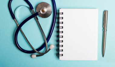 Stetoskop, boş bir not defteri ve açık mavi arka planda duran bir kalem. Üst görünüm kapat