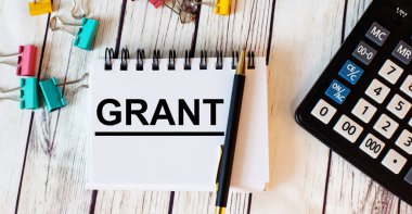 Açık ahşap bir masada hesap makinesi, çok renkli ataçlar ve kalemli bir defter ve GRANT