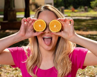 Mutlu ve gülümseyen genç bir kadın turuncu bir kesikle gözlerinin önünde oynuyor. Fotoğraf açık hava parkında ve ağaçların altında