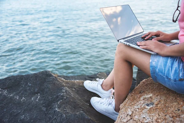 Young Woman Freelance in pink shirt sitting on the rock and working with laptop at outdoor. Sea background.