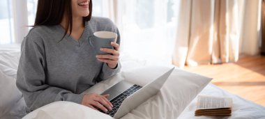 Gülümseyen bir fincan çay tutarken dizüstü kullanarak gri süveterli Asyalı güzel kadın. Rahatlatıcı tatil kavramı. Kopya alanı.