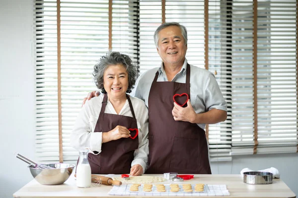 Happy Asian senior couple in brown aprons are holding red cookies cutters in heart shape over their chest. Looking at the camera.