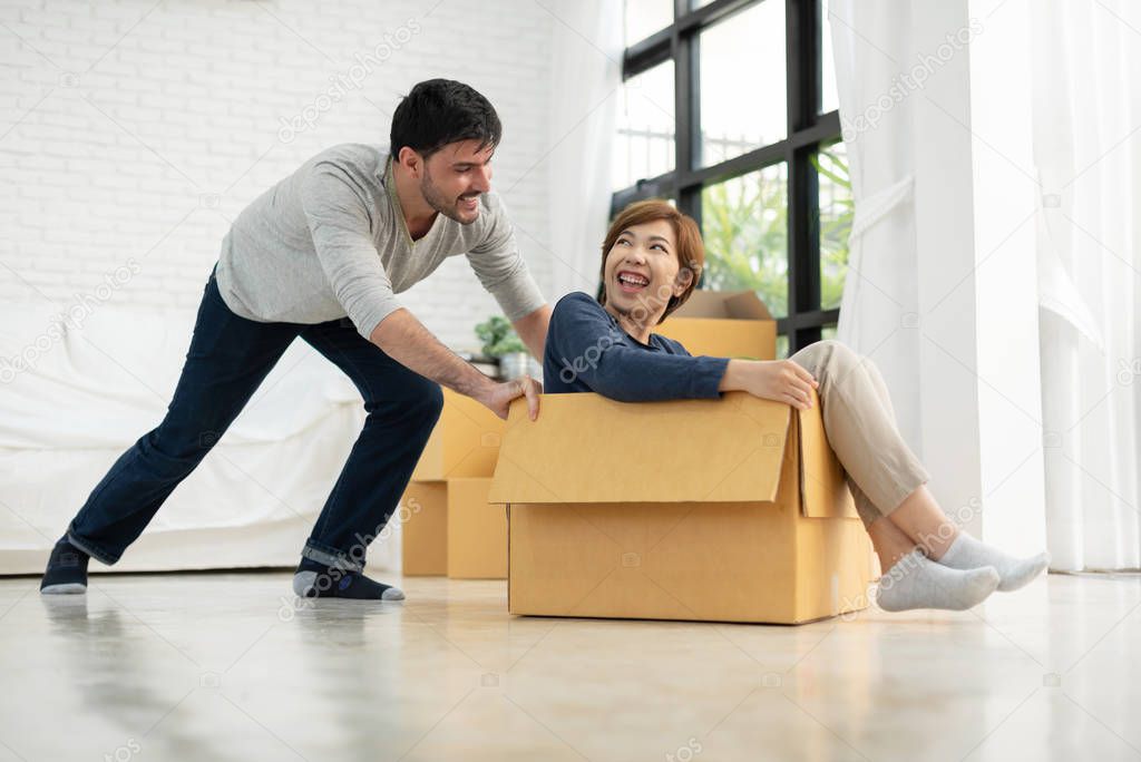 Happy Young couple having fun with cardboard boxes in new home at a moving day. Man and Woman playing with carton box. Multiethnic people enjoying.
