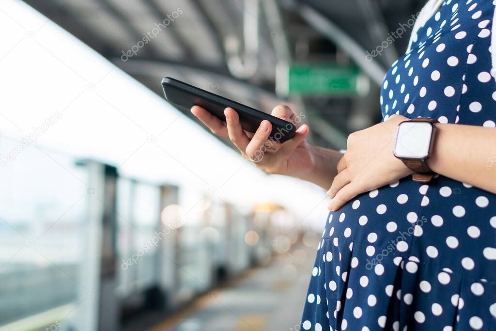 Pregnant woman enjoying internet technology while using mobile phone or smartphone during waiting commuter bus or train for traveling to work in the city. 