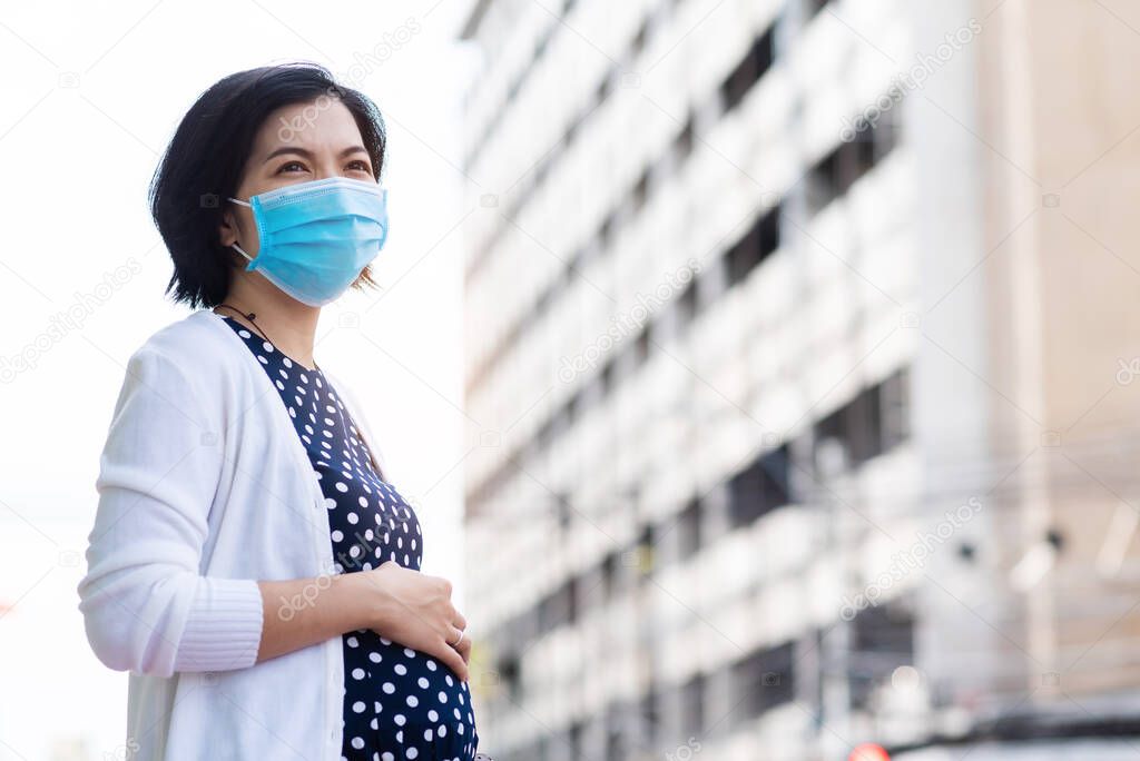 Worried Asian pregnant woman in protective face mask for air pollution and pandemic coronavirus or covid-19 touching her belly and standing against building background at outdoor with new normal life