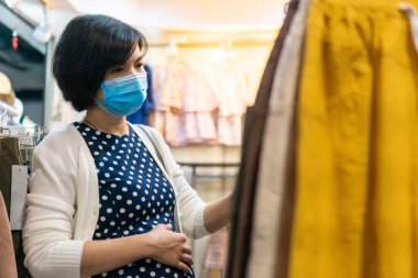 Asyalı hamile bir kadın butikte yüz maskesi takıyor, genç bir kadın bir mağazada bebek kıyafetleri seçiyor ve bakıyor..