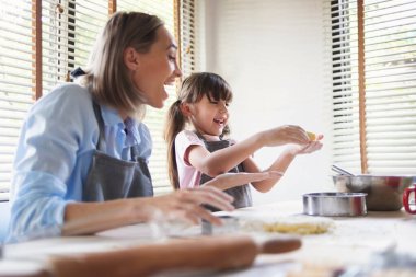 Anne ve küçük kızı evde kurabiye pişirirken gülüp eğleniyorlar. Gülümseyen aile pasta yaparken eğleniyor. Mutlu kadın ve çocuk birlikte masaya un serpmece oynuyorlar..