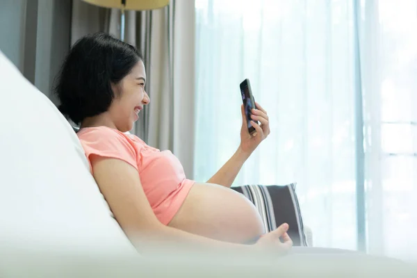 Attractive Asian Pregnant woman in pink shirt making video call online for talking with family on smartphone. Happy smiling Young Mother using mobile phone while pregnancy. Internet and communication