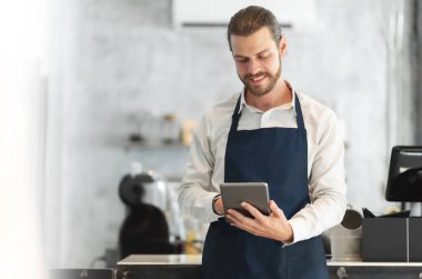 Modern kafelerin barında dijital tablet kullanan mavi önlüklü bir hippi portresi. Gülümseyen çekici sakallı adam internetten sıcak içecek siparişi alıyor.