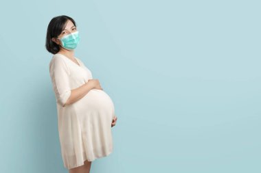 Mutlu gülümseyen Asyalı hamile kadın koruyucu yüz maskesi takıyor mavi izole arka planda duruyor. Kendinden emin, genç, beklenen bir kadın. Kopya boşluğuyla karnına dokunuyor..