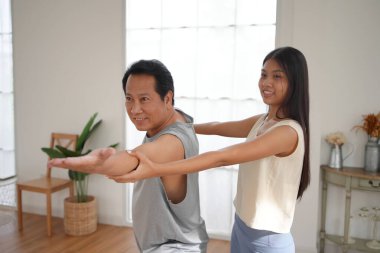 Orta yaşlı Asyalı adam evde yoga yaparken gülümserken, genç bayan antrenörü kollarını esnetirken ona yardım ediyor..