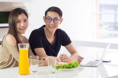 Asyalı genç çift mutfakta oturuyor ve kameraya bakıyor, yanında iki bardak taze portakal suyu, sebze salatası, dizüstü bilgisayar ve akıllı telefon var. Mutlu bir çift.