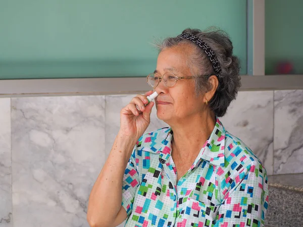 Asian elderly woman using herbal inhaler while sitting on chair