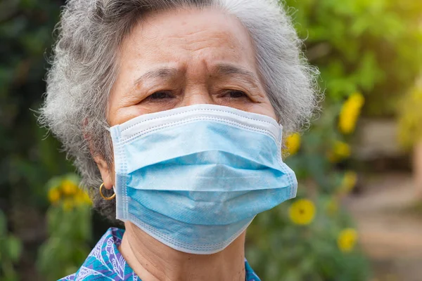A portrait of an elderly woman wearing a face mask looking away while standing in a garden. Mask for protect virus, coronavirus, pollen grains. Concept of old people and healthcare.
