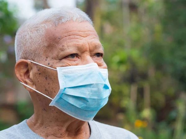 A portrait of an elderly man wearing a face mask looking away while standing in a garden. Mask for protect virus, coronavirus, pollen grains. Concept of old people and healthcare