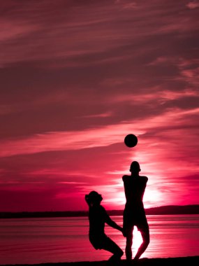Yatay renkli bir resim. İkiz silüetleri olan bir adam ve bir kadın akşam deniz kenarında kızıl gün batımına karşı voleybol oynuyorlar.
