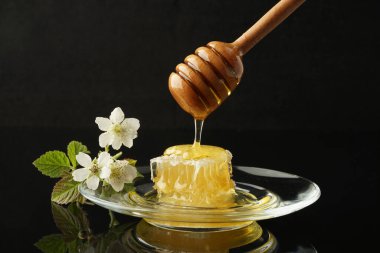 Siyah arka planda çiçeklerle şeffaf bir plakanın üzerinde duran, kaşıktan bal damlayan sıvı çiçekleri taraklar.