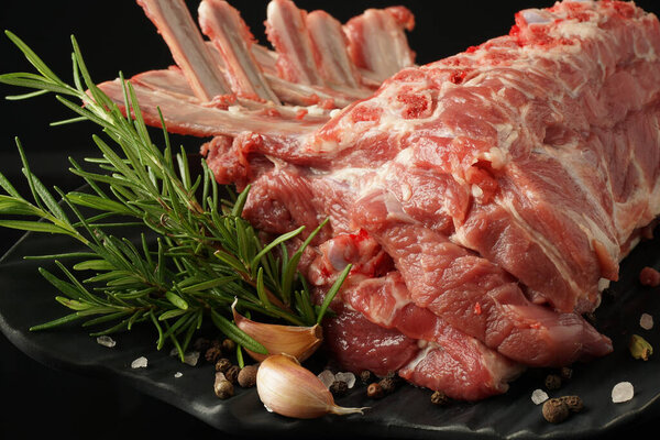 свежая стойка ягненка на черной тарелке на темном фоне со специями: розмарин, перец, соль, чеснок