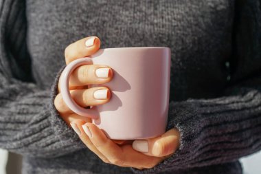 Güzel, bakımlı beyaz manikürlü kadın elleri bir bardak sıcak içecek tutuyor. Ellerin pozisyonu, kalın örülmüş gri bir süveterin arka planına karşı ön taraftadır.