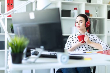 Genç bir kız kulaklık ofiste bir masada oturuyor, kırmızı bir fincan elinde tutan ve ekrana görünüyor.