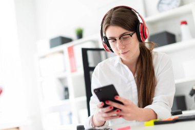 Genç bir kız kulaklık ofiste bir masada oturur ve bir telefon elinde tutan.