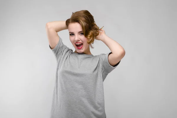 Uroczy młoda dziewczyna w szary T-shirt na szarym tle. Dziewczyna trzyma ręce za głową i krzyki — Zdjęcie stockowe